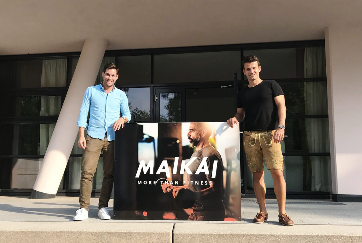 Die Gründer von Maikai stehen vor dem Eingang und halten ein Schild mit der Aufschrift "Maikai – More than fitness".
